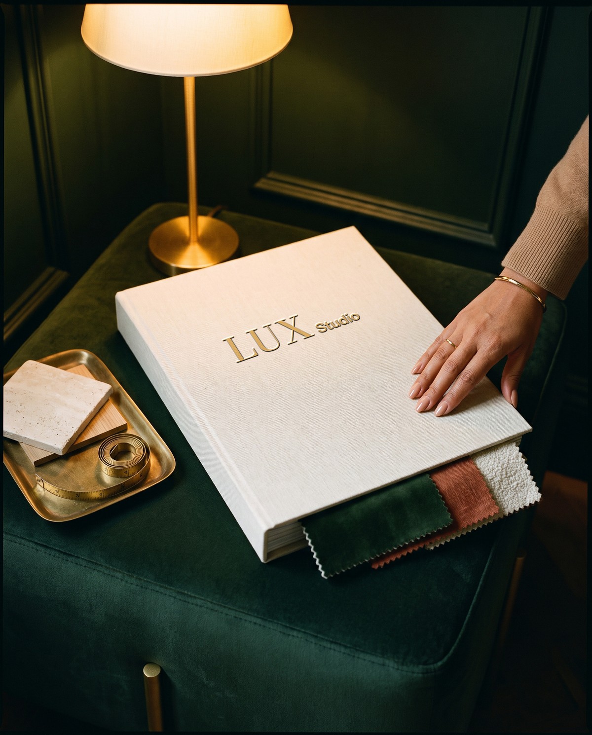 A hand on a linen folio foiled in gold, fabric samples tucked inside, under a lamp